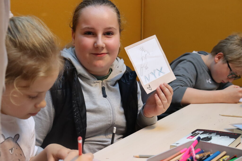 Dziewczynka w szarej bluzie prezentuje rysunek papugi z podpisem: Nyx.