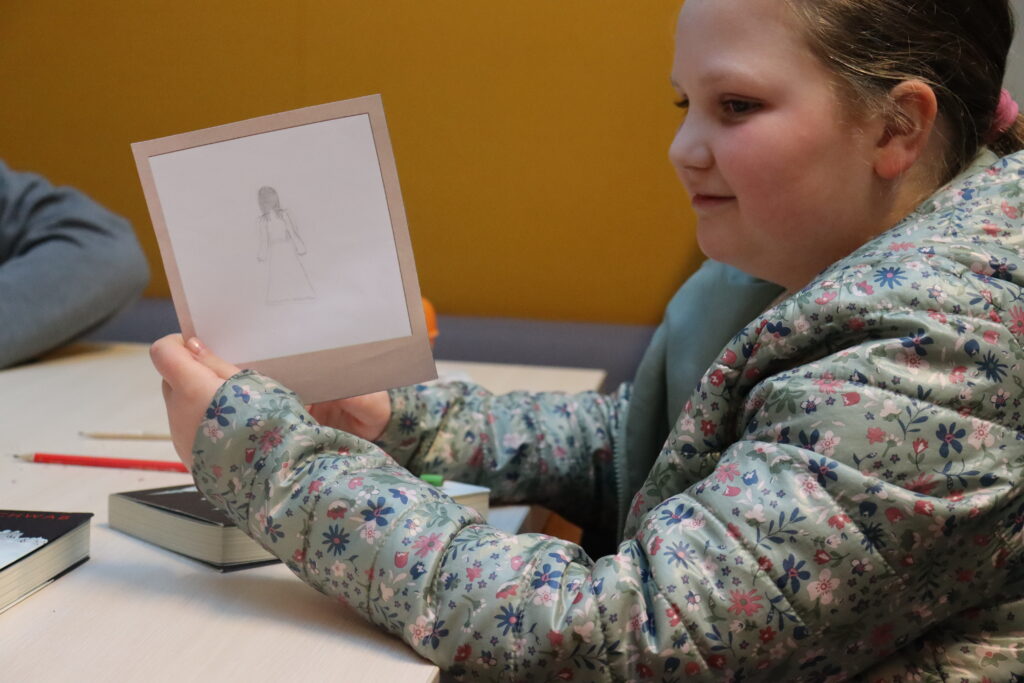 Dziewczynka z kurtce w kwiatki pokazuje swój rysunek: blady duch w długiej sukni.