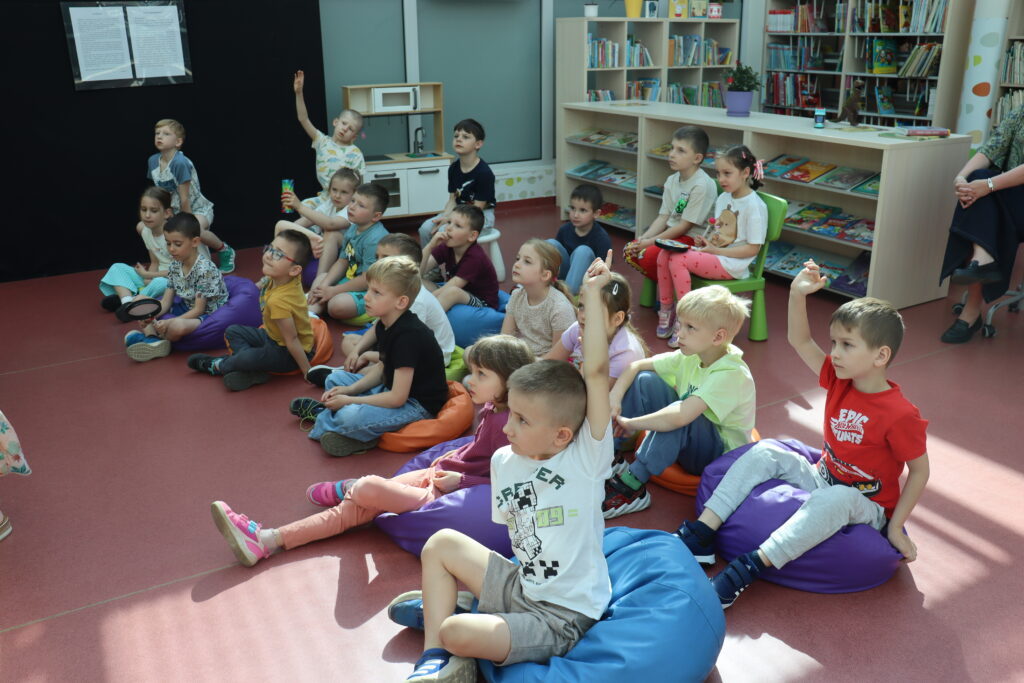 Grupa przedszkolaków siedzi na kolorowych pufach w bibliotece, uważnie słuchając i zgłaszając się do odpowiedzi podczas zajęć.