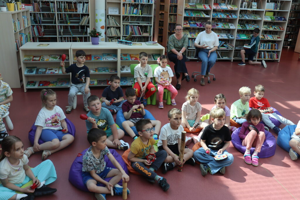Przedszkolaki siedzą na pufach w kole w bibliotece, trzymając małe instrumenty perkusyjne i bębny.