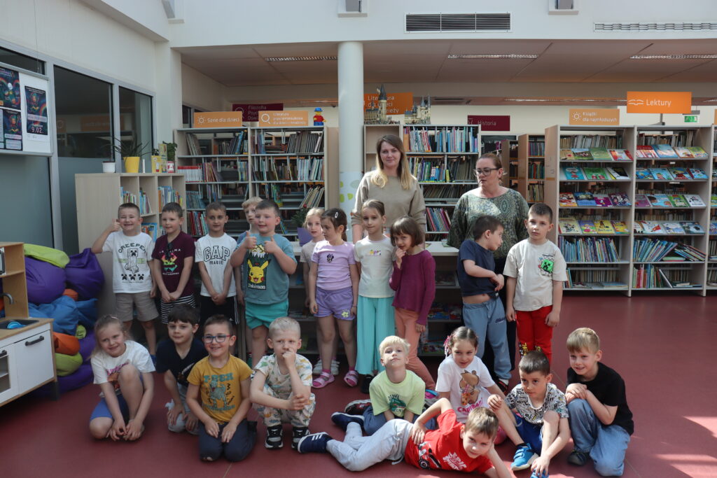 Grupa przedszkolaków i dwie nauczycielki, w tym Marta Grucela, pozują do pamiątkowego zdjęcia między regałami z książkami w bibliotece.